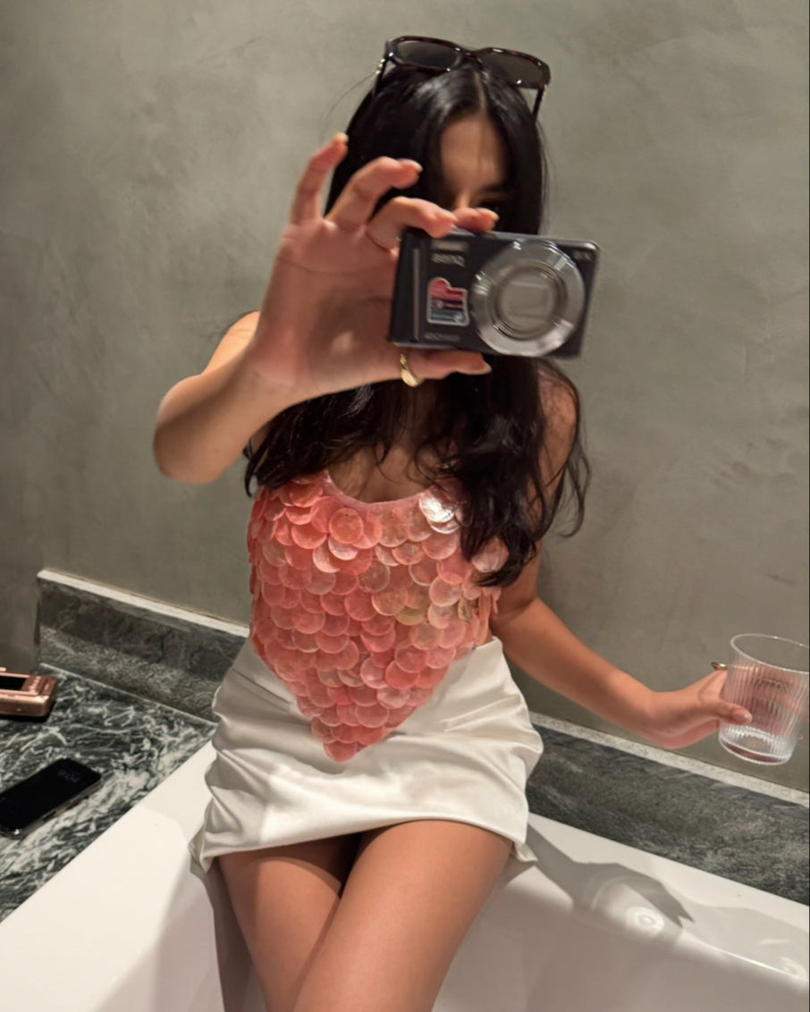 Person taking a mirror selfie in a bathroom with a pink shell top and white skirt.