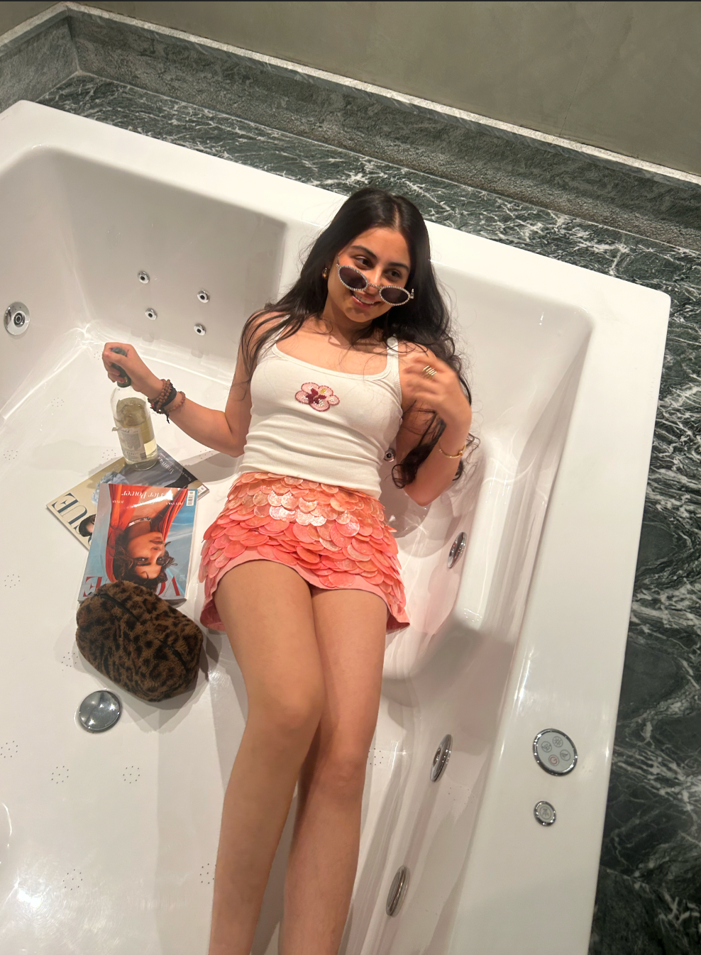 Woman in a bathtub holding a bottle and a magazine, with a leopard print bag beside her.